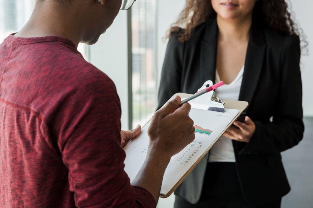 two people having a conversation, one holding a clipboard with paper on it