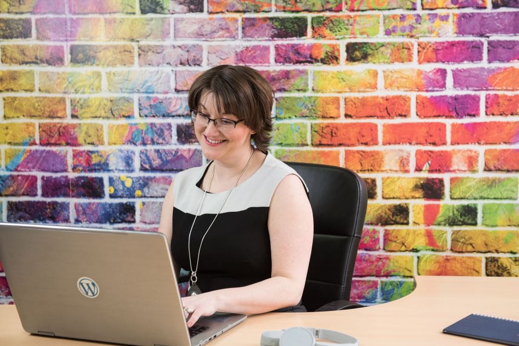 Elizabeth sitting by a colourful wall using a laptop