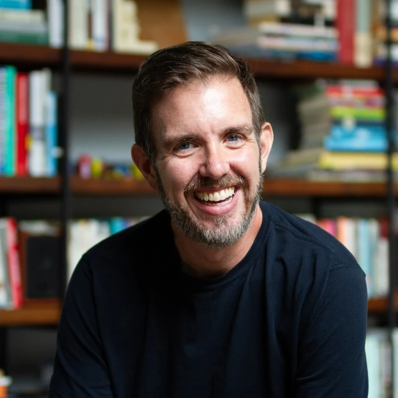 Brett Harned with a beard, next to a shelf of books, wearing a navy top.